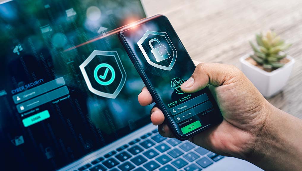 A person holding a smartphone displaying a cybersecurity login screen with a fingerprint scanner in front of a laptop